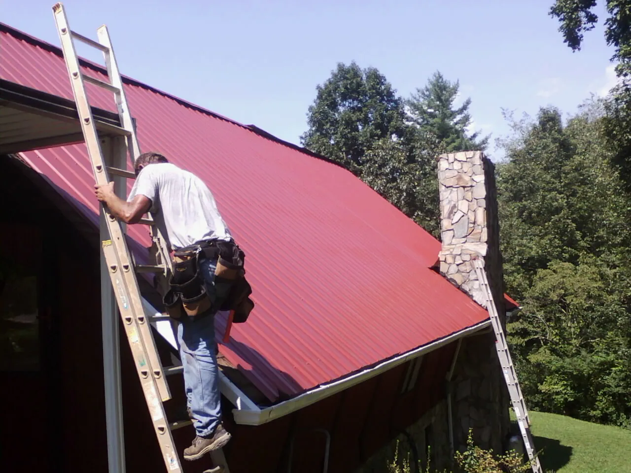 Roofer on a ladder beside a red metal roof and stone chimney — Soffit & Fascia Repair by Roofing Brothers in Meridian